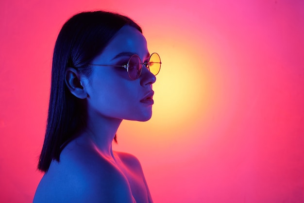 Luz azul que cae sobre una chica desnuda seria en elegantes gafas redondas sobre fondo rosa brillante