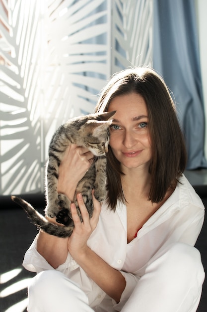 À luz do sol, uma jovem de camisa branca segura um gatinho cinza nos braços