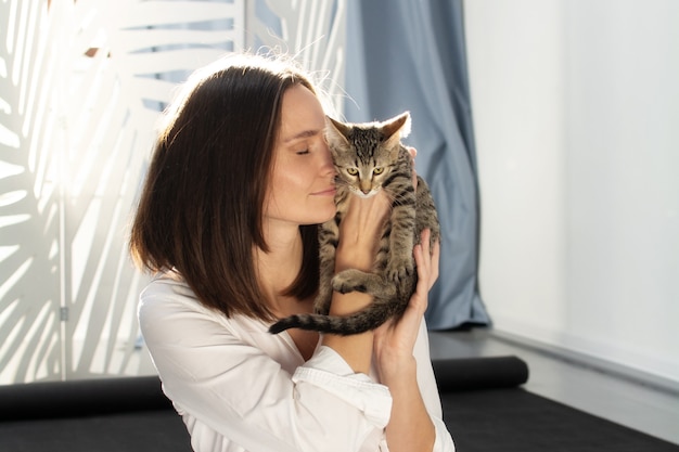 A la luz del sol, una mujer joven con una camisa blanca sostiene un gatito gris en sus brazos