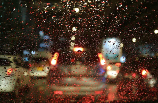 Luzes da rua turva e lanternas traseiras vistas através das gotas de chuva no pára-brisas do carro à noite