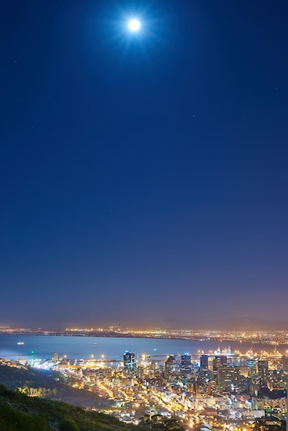 Luzes urbanas da cidade com lua cheia no céu da meia-noite com espaço de cópia Skyline com iluminação colorida com o oceano aberto no horizonte Edifícios da cidade à noite em Signal Hill África do Sul