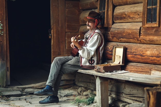 Lviv Ucrania 14 de octubre de 2019 Hutsul en traje nacional toca la guitarra mientras se sienta cerca de su casa de troncos