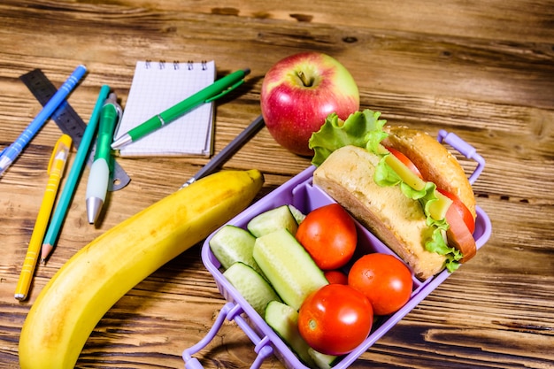 Maçã madura, banana, artigos de papelaria diferentes e lancheira com hambúrguer, pepino e tomate na mesa de madeira rústica