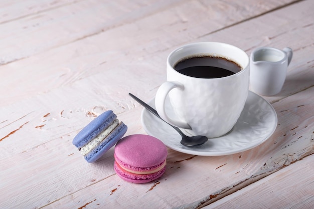 Macaroons brilhantes e uma xícara de café branca em uma mesa de madeira clara