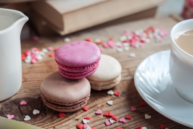 Macaroons e uma xícara de café um jarro de leite em um fundo de pequenos corações em uma vista superior de fundo de madeira