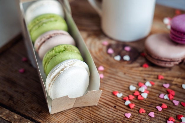 Macaroons em uma caixa uma xícara de café um jarro de leite no contexto de pequenos corações em uma vista superior de fundo de madeira