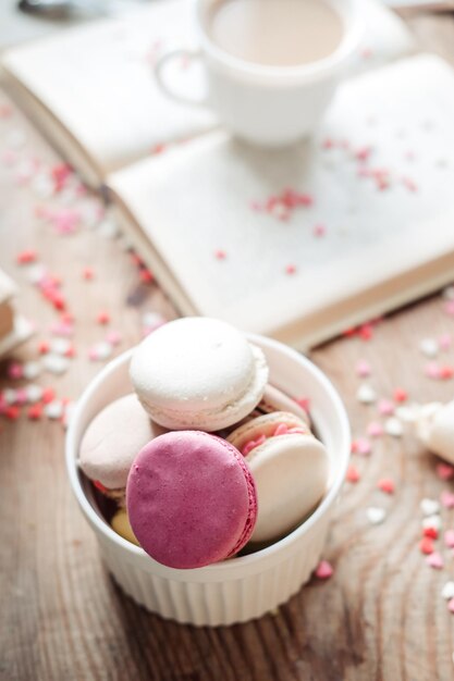 Macaroons em uma tigela e uma xícara de café um livro sobre o fundo de pequenos corações em uma vista superior de fundo de madeira