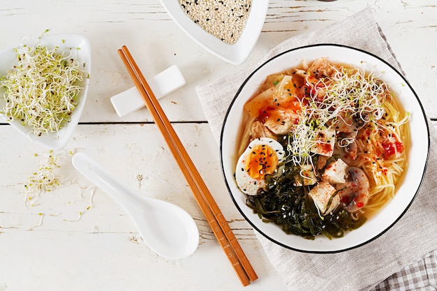 Macarrão asiático do ramen do miso com kimchi do repolho, alga, ovo, cogumelos e tofu do queijo na bacia na tabela de madeira branca. cozinha coreana. vista do topo. lay plana