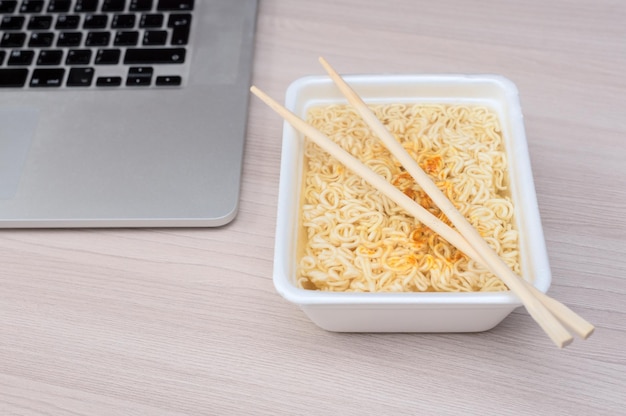 Macarrão chinês instantâneo em embalagens para viagem com palitos de madeira e laptop na mesa de escritório Foco seletivo Comida asiática e conceito de entrega Alimentação pouco saudável