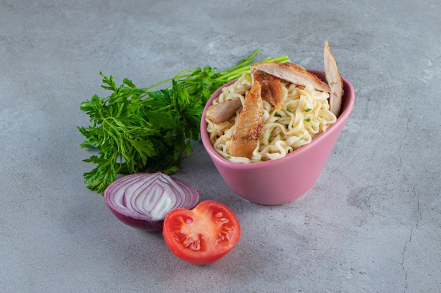 Macarrão com carne em uma tigela ao lado do ramo de salsa, tomate e cebola, na superfície de mármore.