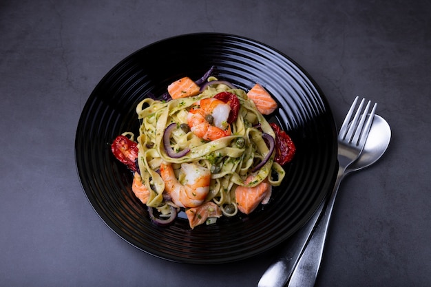 Macarrão com frutos do mar, tomates secos, alcaparras e cebolas roxas. Massa caseira com camarão, salmão (truta) e molho pesto. Fundo preto, placa preta. Fechar-se.