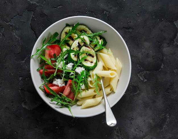 Macarrão de almoço vegetariano e salada de rúcula feta de tomate de abobrinha grelhada em uma vista superior de fundo escuro