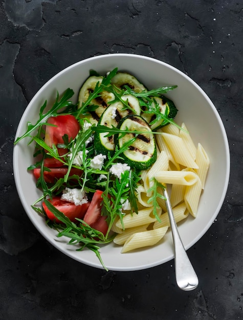 Macarrão de almoço vegetariano e salada de rúcula feta de tomate de abobrinha grelhada em uma vista superior de fundo escuro