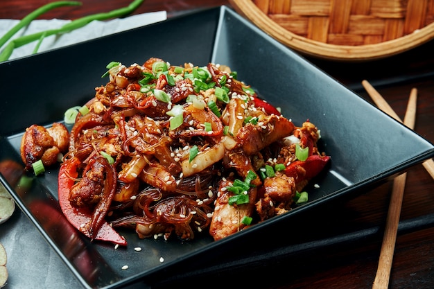 Macarrão de arroz com milho bebê, pimentão, cebola, cenoura e frango frito em um wok com molho teriyaki em chapa preta na mesa de madeira. saborosa comida de rua asiática