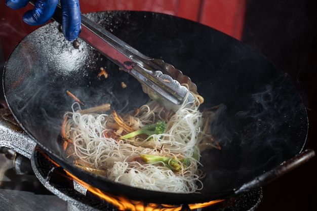 Macarrão de arroz Funchoza com legumes cozinhando no fogo na frigideira wok. Comida flambeada de rua.