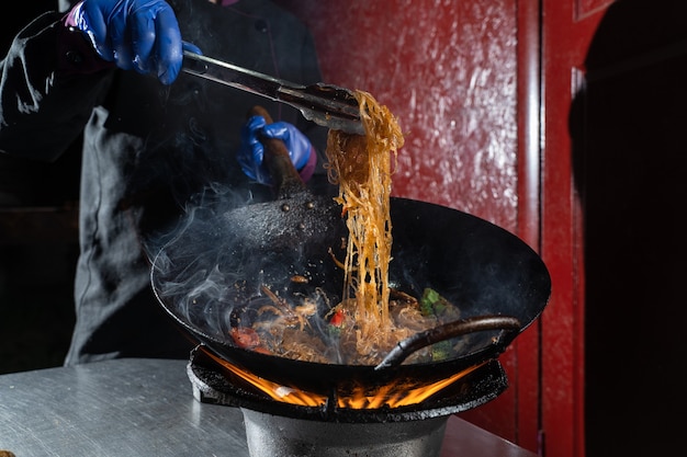 Macarrão de arroz Funchoza com legumes cozinhando no fogo na frigideira wok. Comida flambeada de rua.