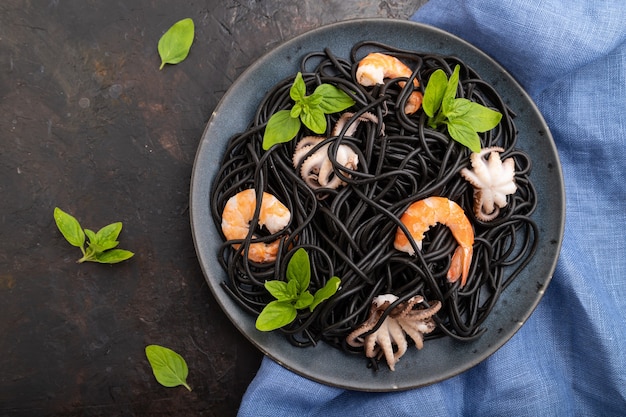 Macarrão de tinta de choco preto com camarões ou camarões e pequenos polvos em fundo de concreto preto e tecido azul. Vista superior, configuração plana,