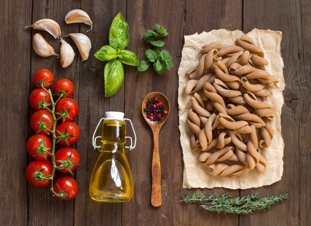 Foto macarrão de trigo integral, legumes, ervas e azeite na vista superior da mesa de madeira