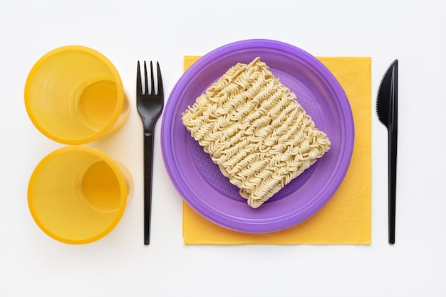 Macarrão em um prato de plástico lilás, vidro amarelo, garfo e faca pretos, vista superior.