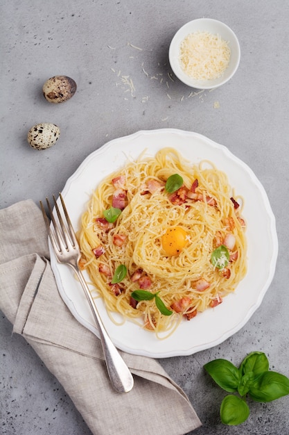 Macarrão espaguete à carbonara com bacon, queijo parmesão, gema de ovo e folhas de manjericão na superfície cinza clara. Prato tradicional italiano