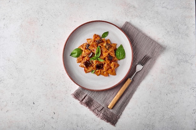 Macarrão farfalle com berinjela, molho de tomate e manjericão no prato com garfo e guardanapo no fundo cinza