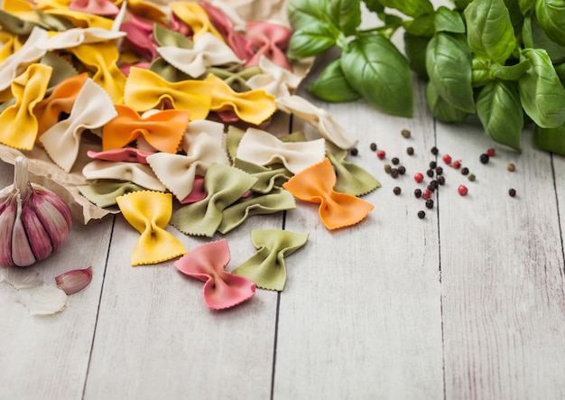 Macarrão farfalle tricolor cru em papel pardo sobre fundo de mesa de madeira clara com manjericão e alho