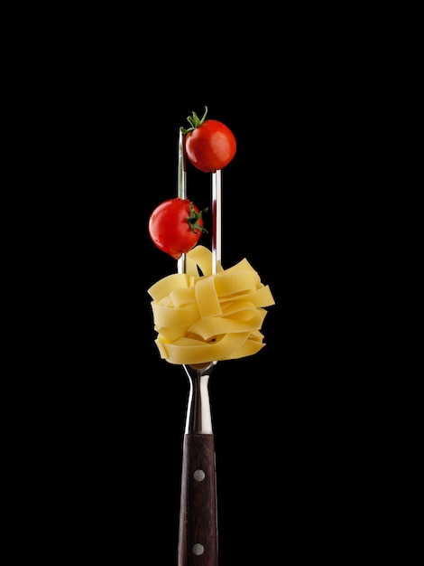 Macarrão fettuccine cru com tomate cereja em um garfo de duas pontas. Fundo preto, isolado.