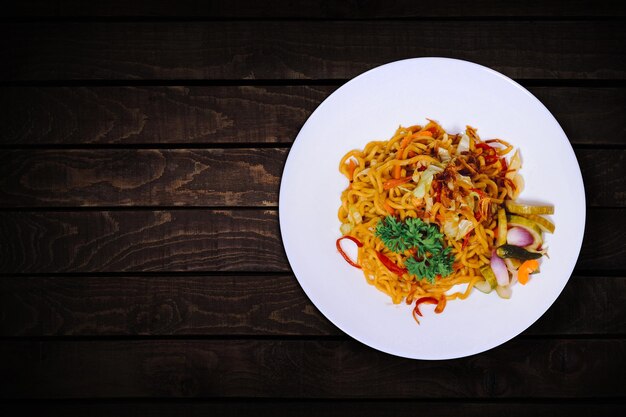 Macarrão frito picante com várias adições na chapa branca na vista superior da mesa de madeira escura