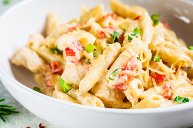 Macarrão penne com frango, pimenta e cebolinha em molho cremoso em um prato branco, fundo cinza.