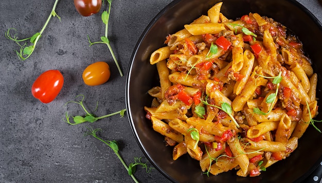 Macarrão penne em molho de tomate com carne, tomate decorado com brotos de ervilha em uma mesa escura. vista do topo