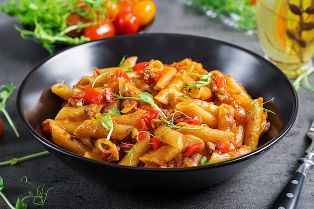 Macarrão penne em molho de tomate com carne, tomate decorado com brotos de ervilha em uma mesa escura.