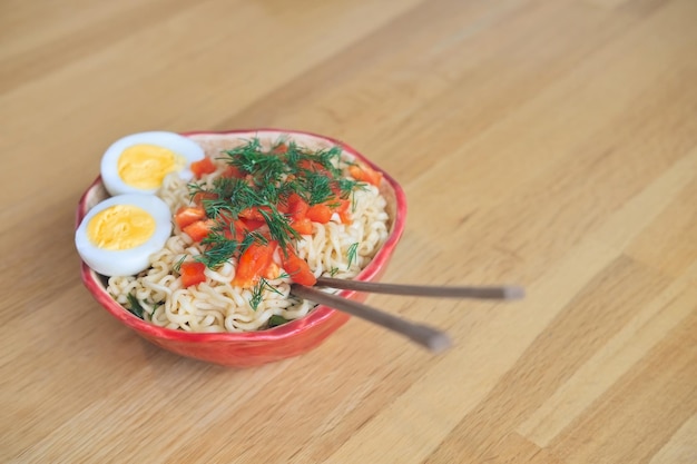 Macarrão ramen com ovo, ervas e legumes em uma tigela vermelha com pauzinhos em uma mesa de madeira