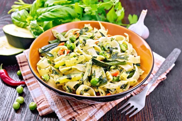 Macarrão tagliatelle com abobrinha, ervilha, feijão de espargos, pimenta e espinafre em um prato na toalha, alho, garfo e manjericão no fundo de uma placa de madeira escura