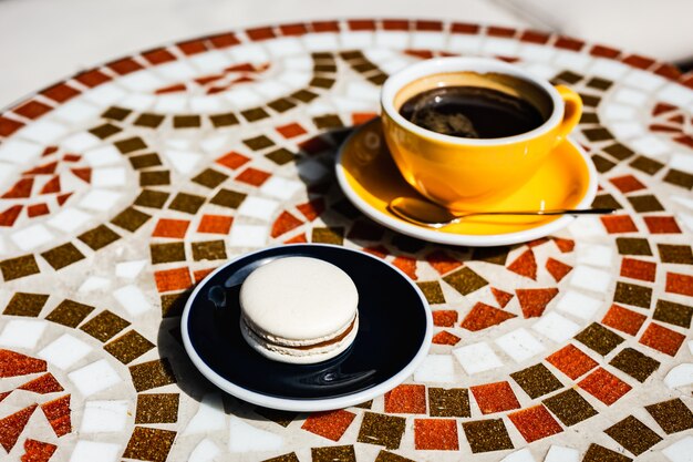 Macarrón de vainilla con chocolate en un plato, una taza amarilla de café negro, en una mesa de piedra de mosaico de un café en un día soleado