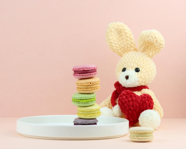 Macarrones coloridos en plato blanco sobre fondo rosa claro con un conejo hecho a mano con corazón rojo