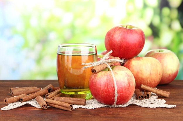 Maçãs maduras com paus de canela e copo de bebida de maçã na mesa de madeira sobre fundo brilhante