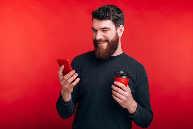 Machen Sie eine Pause, lesen Sie Neuigkeiten Ein lächelnder bärtiger Mann schaut auf sein Handy und hält einen roten Pappbecher in der Hand.