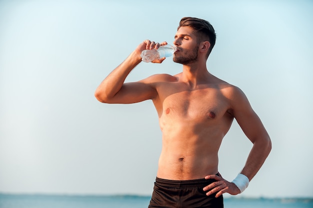 Machen Sie eine Pause zum Trinken. Selbstbewusster junger muskulöser Mann, der Wasser trinkt und die Hand an der Hüfte hält, während er im Freien steht