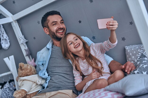 Machen Sie tolle Erinnerungen. Junger Vater und seine kleine Tochter machen Selfie, während sie zu Hause auf dem Bett sitzen