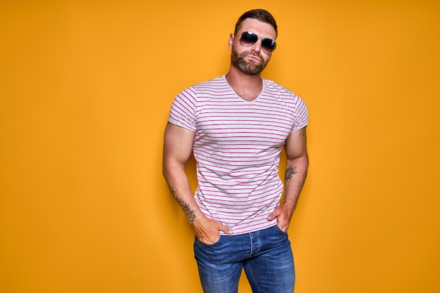 Macho barbudo confiante musculoso em camisa jeans e óculos de sol elegantes em pé no estúdio sobre amarelo ...