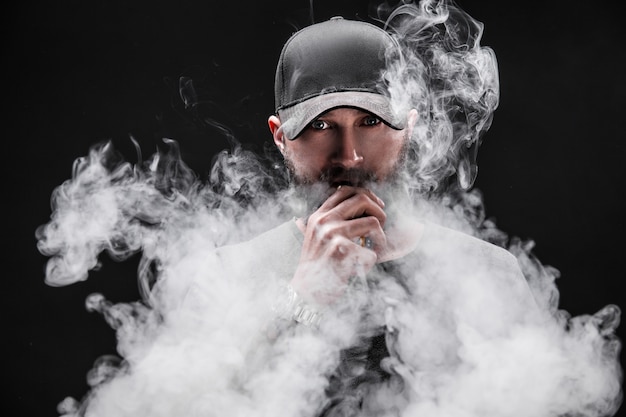 Macho barbudo, vestido com uma camisa cinza, óculos escuros e boné de beisebol vaping