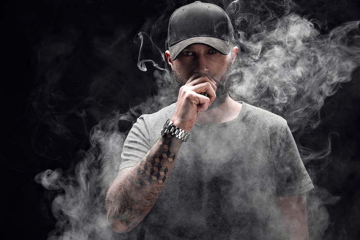 Foto macho barbudo, vestido com uma camisa cinza, óculos escuros e boné de beisebol vaping