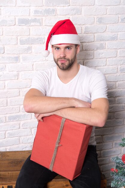 Macho com chapéu de Natal segurar presente embrulhado. Homem santa com caixa vermelha na parede de tijolo branco. Conceito de Boxing Day. Oferta e troca de presentes. Preparação e celebração de feriados.