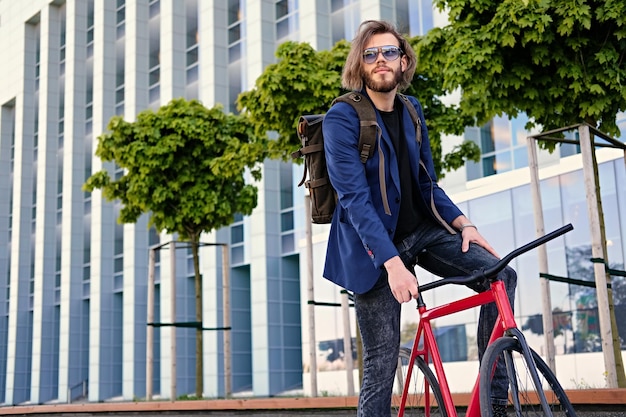 Macho hipster barbudo com mochila senta-se na bicicleta fixa vermelha em um parque.