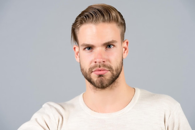 Macho im T-Shirt auf grauem Hintergrund, Mode. Mann mit bärtigem Gesicht, stilvollem Haar, Haarschnitt, Salon. Mode, Stil, Trend. Friseursalon, Friseursalon. Pflege, männliches Schönheitskonzept