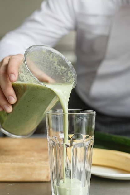 Macho jovem em uniforme derramando smoothie fresco de cor verde em vidro limpo