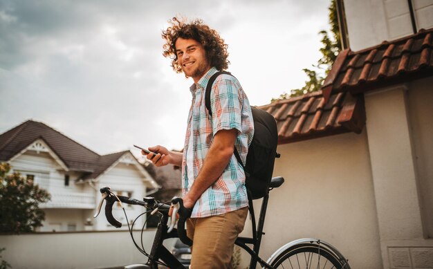 Foto macho jovem hippie com cabelo encaracolado, olhando para a câmera andando na rua da cidade usando seu celular para aplicação gps feliz sorridente cara ciclista com mochila navegando na internet usando telefone inteligente