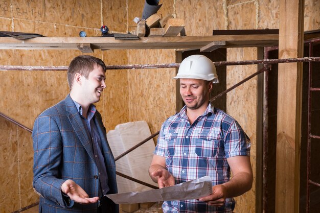 Macho joven arquitecto y capataz de trabajador de la construcción inspeccionando planes de construcción juntos dentro del edificio inacabado