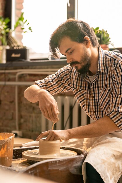 Foto macho joven artesano inclinado sobre la pieza de arcilla giratoria mientras le da forma a la rueda en el taller