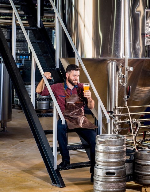 Macho joven cervecero en delantal de cuero en la fábrica de cerveza moderna